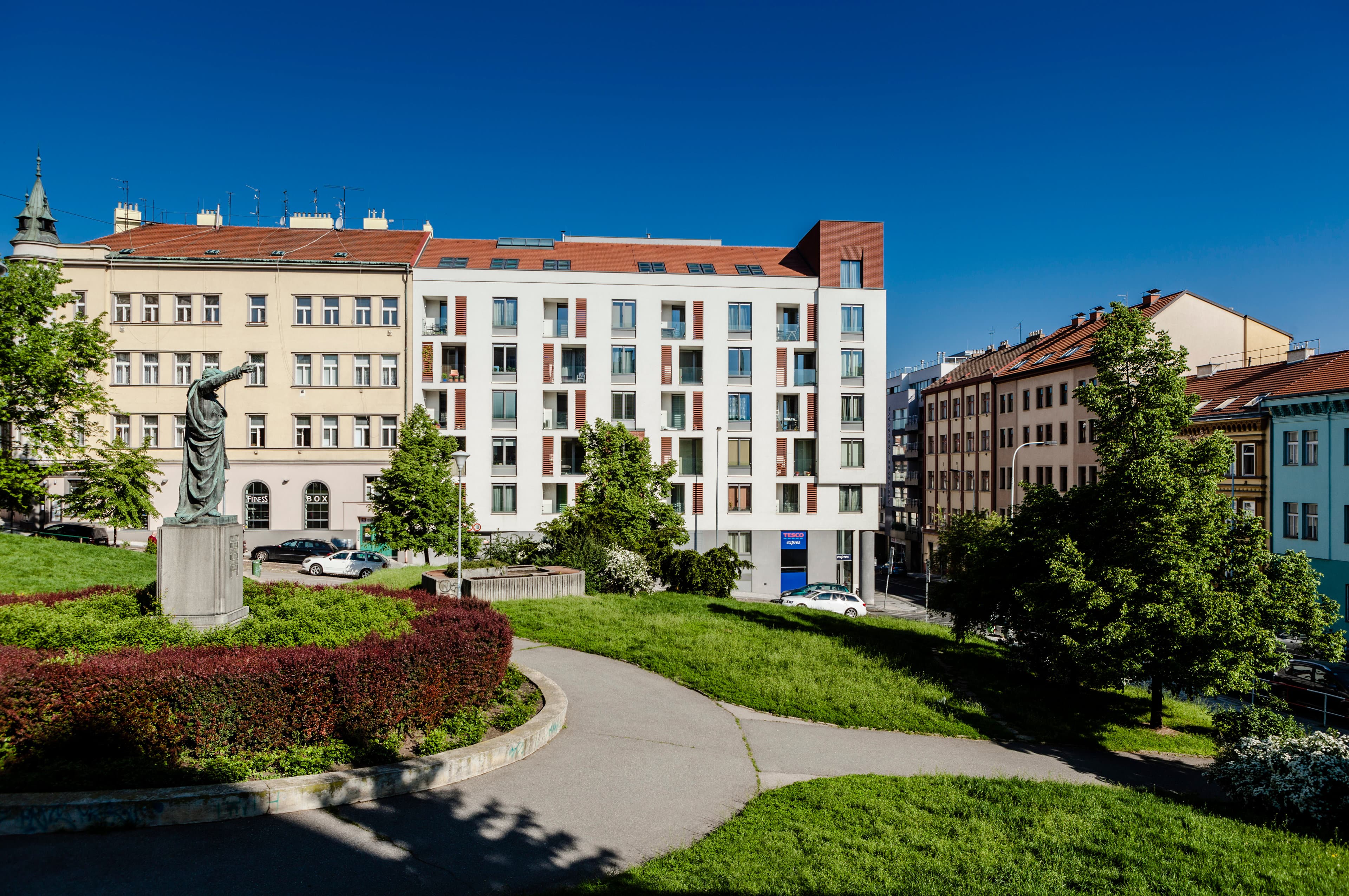 Bytový dům na severozápadní straně Havlíčkova náměstí, pohled z parku přes zeleň – bílá fasáda a šikmá střecha mezi staršími činžovními domy