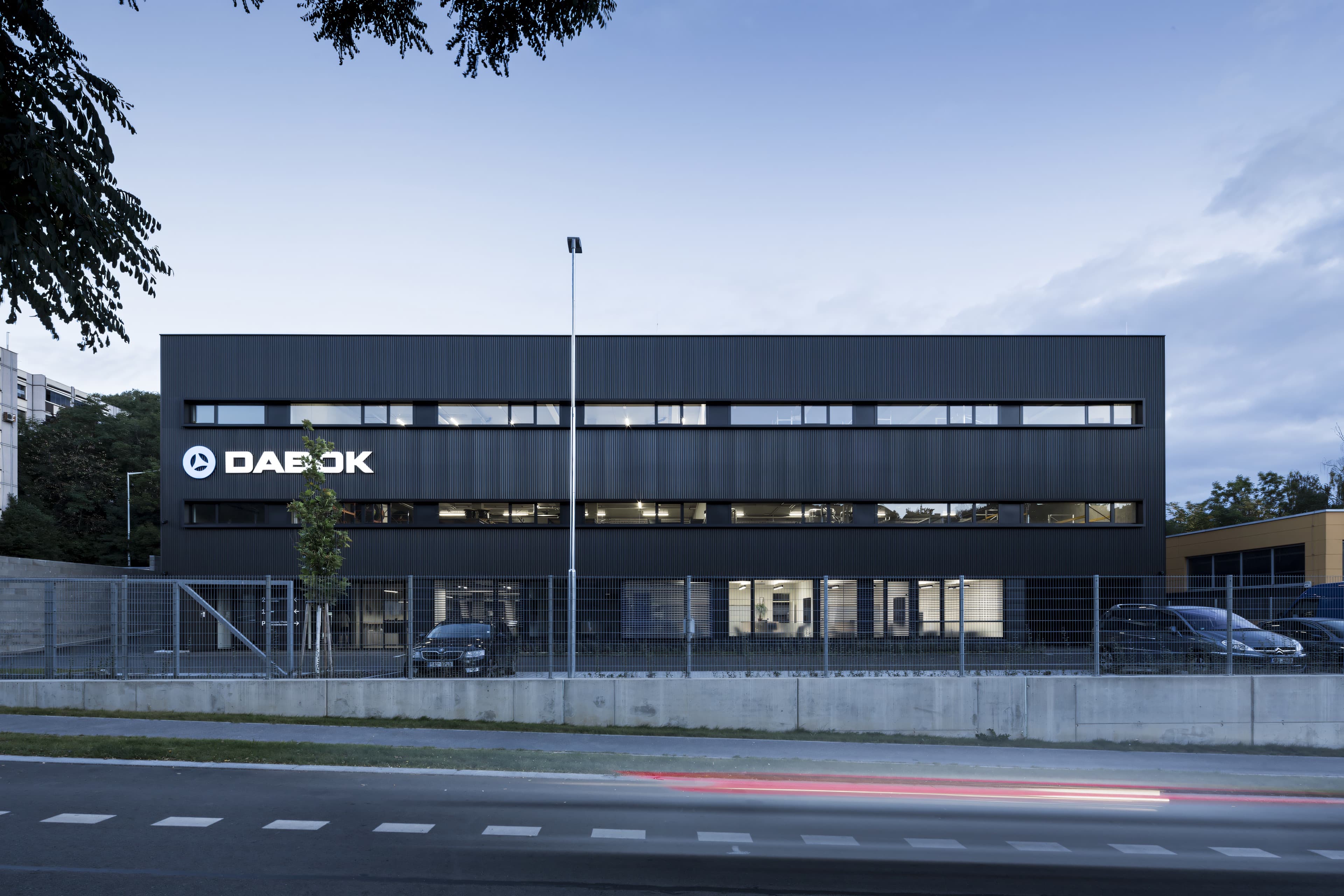 Sklad DABOK po setmění, pohled z ulice Ke Kotlářce – černá plechová fasáda, prosvětlená okna showroomu a nasvícené logo firmy