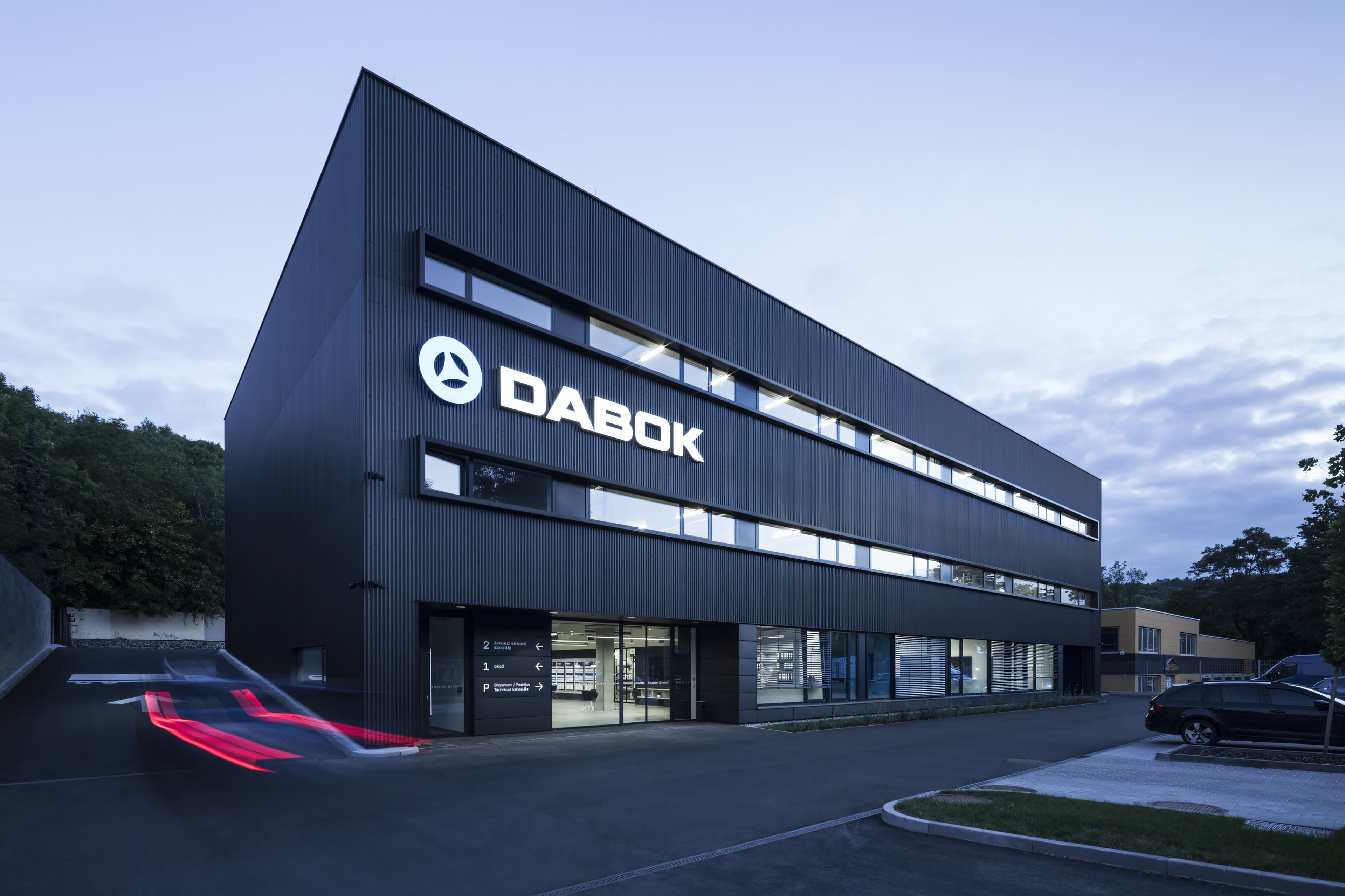Budova od vjezdu do areálu po setmění – černá hmota s nasvíceným logem, ve spodním podlaží prosvětlený showroom